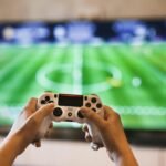 E-sports em Expansão: Os Jogos Competitivos Que Estão em Alta Agora Close-up of hands holding a game controller playing a football video game on screen.